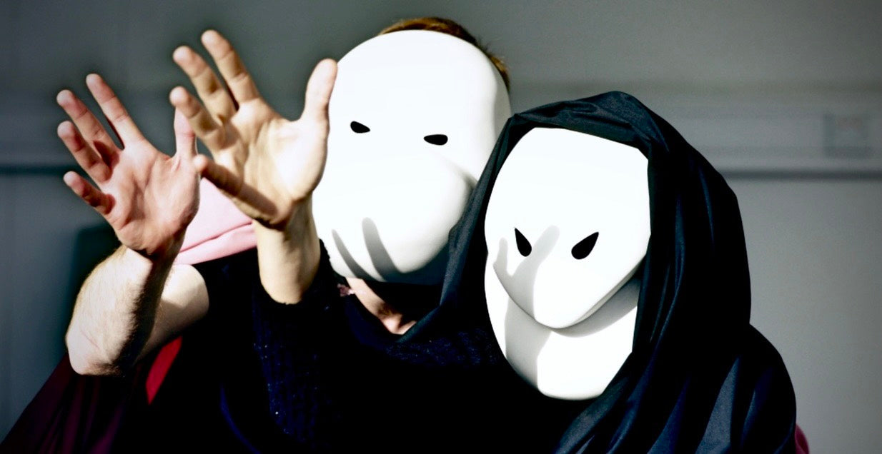 Two people wearing white masks with black eyes against a neutral background